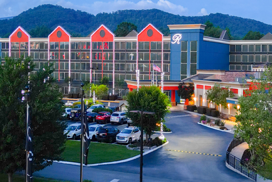 Hotel building with a glass facade and red accents, surrounded by trees and hills, with a parking lot filled with cars in front.