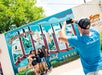 A man in a blue shirt takes a photo of four women posing in front of a colorful "Greetings from Austin, Texas" mural on a sunny day.