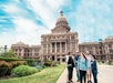Five people walk together on a path in front of a large, historic capitol building with a dome under a partly cloudy sky.