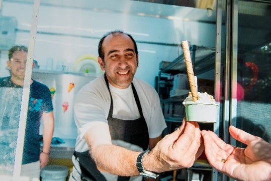 A man wearing an apron hands a cup of ice cream with a wafer stick to a customer through a service window.