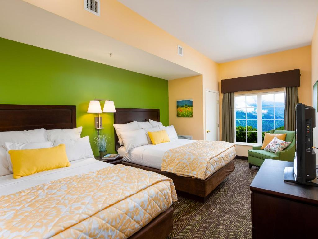 A hotel room with two double beds, green accent wall, and yellow bedding. A window shows a mountain view. There's a small green sofa and a flat-screen TV on a wooden stand.