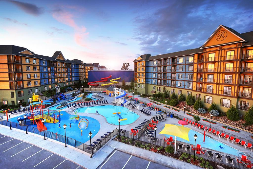 Aerial view of a resort featuring a large outdoor pool with slides, surrounded by multi-story buildings and lounge chairs, under a colorful sky at sunset.