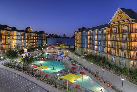 A hotel with a large outdoor pool area featuring multiple water slides and colorful umbrellas, surrounded by multi-story buildings during evening.
