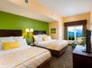 Hotel room with two double beds, green and beige walls, a TV, a window with mountain view, and a small seating area with a green chair and yellow cushions.