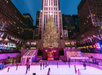 People skating at an ice rink in front of a large illuminated Christmas tree and tall buildings at night.
