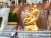 Two people are ice skating near the Prometheus statue at Rockefeller Center, with onlookers behind a railing above and a fountain in the background.