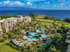 Aerial view of a coastal resort featuring a large pool, palm trees, and buildings, with the ocean and mountains in the background under a partly cloudy sky.