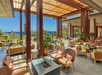 Open-air lounge with wicker seating, potted plants, and a view of the ocean through wooden beams and large columns.