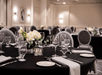 A banquet room full of black cloth covered tables set with plates and glasses and flowers in the middle.