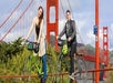 Two people posing with bicycles on a railing, with the Golden Gate Bridge in the background on a clear day.
