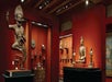 A museum gallery with red walls featuring various Asian sculptures, including a large dynamic figure on the left and a seated Buddha on the right, with several smaller artifacts displayed.
