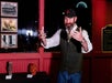 A man with a beard and a cap gestures while speaking indoors, standing beside a display table with small objects and historical photos on the wall behind him.