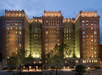 Front view of a large, illuminated historic hotel with an ornate brick facade, surrounded by trees and flanked by additional city buildings under a dusky sky.