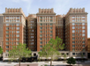 A large, multi-story brick building with intricate architectural details and tall windows, surrounded by green trees, set against a clear sky.
