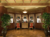 Elegant room with a wooden sideboard, two red chairs, large potted plants, and ornate curtains covering windows. The ceiling features decorative moldings and a central chandelier.