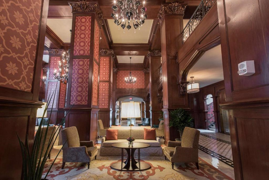 A hotel lobby with vintage decor, featuring ornate wood paneling, red patterned wallpaper, plush seating, and a chandelier.