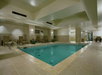 Indoor swimming pool with beige tiled flooring and walls, lounge chairs, potted plants, and overhead lighting.