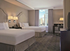 Hotel room with two double beds, a window showing a city view, a desk with a lamp, and a flat-screen TV. Walls are beige with matching carpet and modern lighting fixtures.