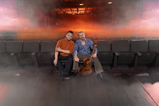 Two people sitting on theater seats with a guitar, surrounded by fog, and an orange glow in the background.