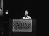 A person stands at a podium speaking into a microphone. The podium displays "The Sound of Simon & Garfunkel" in large letters. A guitar and lamps are visible beside the podium.