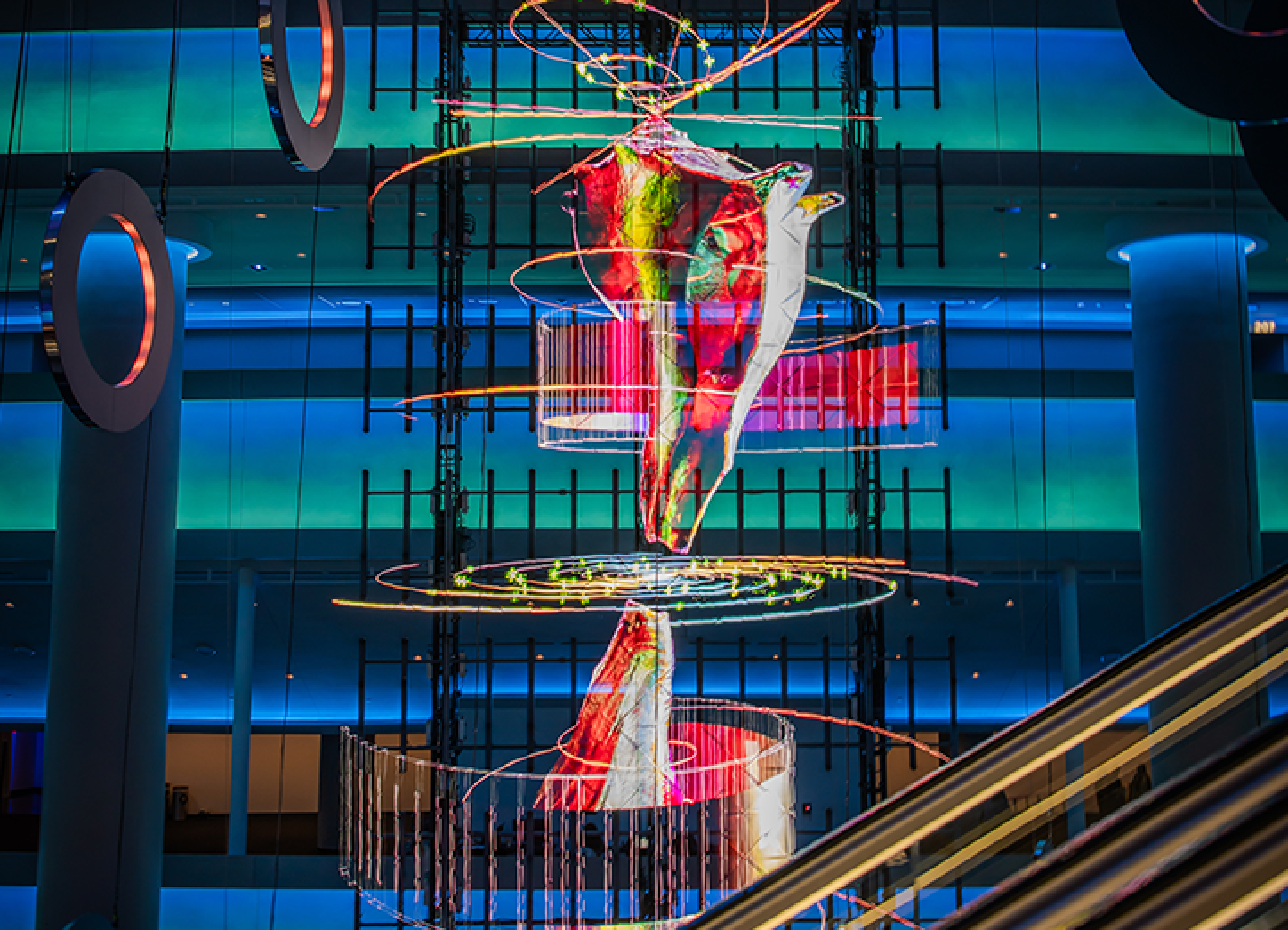 Colorful abstract light sculpture in a multi-story modern building, featuring swirling patterns and vibrant illuminations against a blue background.