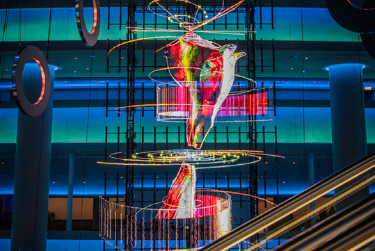 Colorful abstract light sculpture in a multi-story modern building, featuring swirling patterns and vibrant illuminations against a blue background.