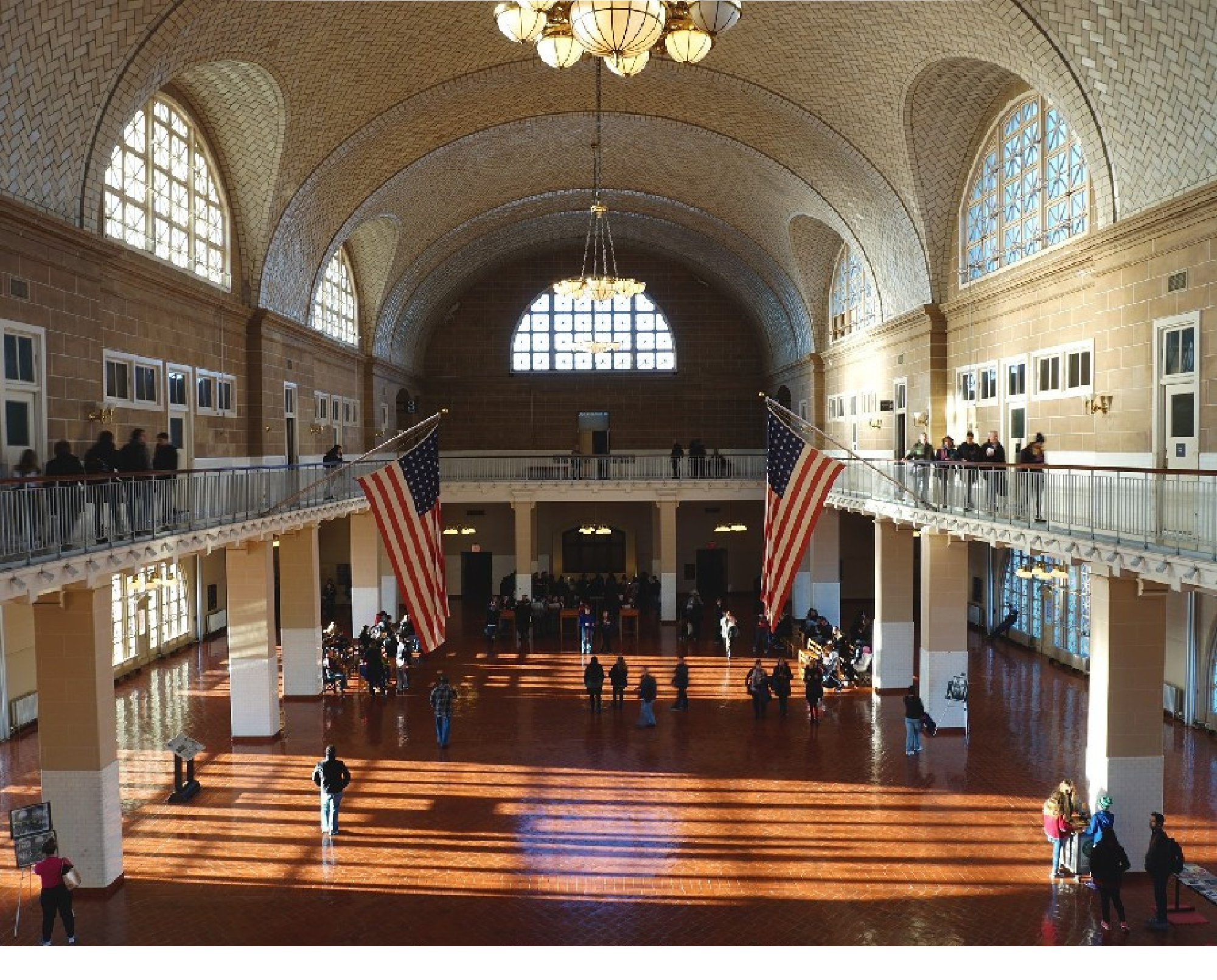 Spacious hall with arched ceiling, large windows, American flags, and visitors. Sunlight casts long shadows on the floor.