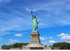 The Statue of Liberty stands on a pedestal against a blue sky with clouds, surrounded by green trees.