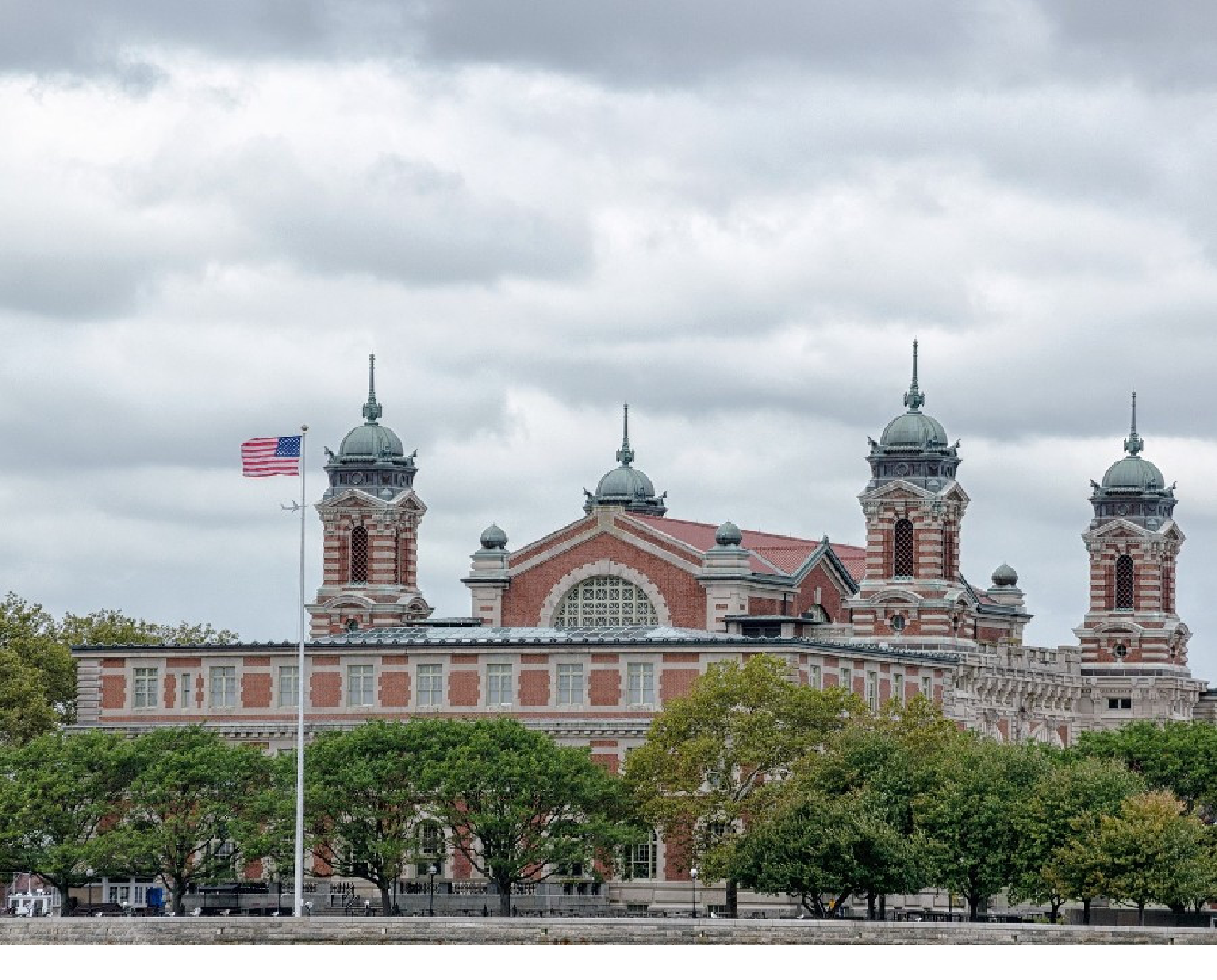 A historic building with ornate architecture, featuring three domed towers, and an American flag in the foreground, set under a cloudy sky.