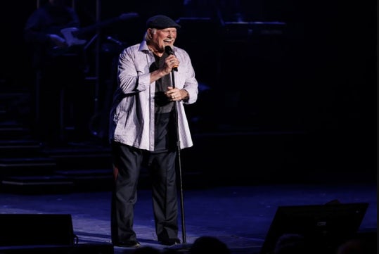 Terry Bradshaw on stage, smiling and singing into a microphone, dressed in a casual outfit with a striped shirt and cap, while a band is visible in the background.