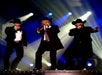 Three men in black suits and cowboy hats sing on stage under dramatic spotlights.