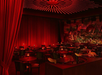 A dimly lit, red-toned room with round tables and chairs. Red curtains and a mural decorate the walls. Warm lighting creates an intimate atmosphere.