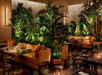 Luxurious restaurant interior featuring tables set with glassware and surrounded by lush green plants. Warm lighting creates an inviting atmosphere.