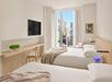 Modern hotel room with two beds, a wall-mounted TV, a desk, and a city view through large windows. Neutral tones with decorative plants and picture frames.