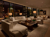 A modern living room with city view at night, featuring white sofas, a coffee table, lamps, and a dining area with a large black pendant light.