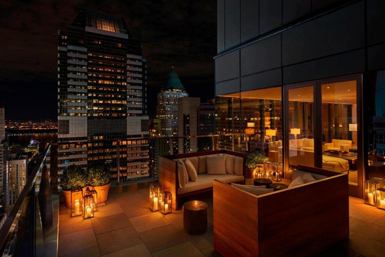 A rooftop terrace at night with seating and lanterns, surrounded by tall city buildings with illuminated windows.