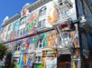 Colorful mural depicting diverse cultural figures on the facade of a multi-story building with fire escapes and a one-way street sign.