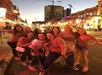 A group of nine women pose and smile together on a brightly lit city street at dusk, with buildings and construction cranes in the background.