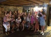 A group of thirteen women pose indoors, holding drinks and smiling at the camera, with wooden floors, exposed beams, and a Jack Daniel's sign in the background.