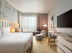 Modern hotel room with a large bed, flat-screen TV, desk, chair, and a seating area. The window has sheer curtains, and the decor is light with a striped accent wall.