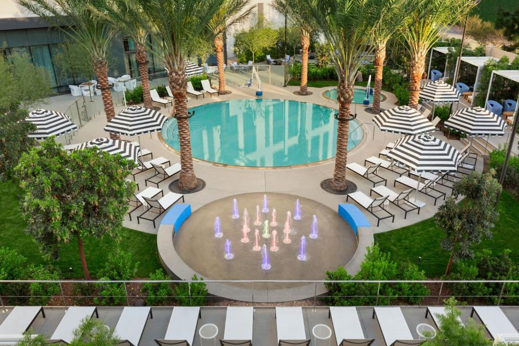 Aerial view of an outdoor circular pool surrounded by lounge chairs and striped umbrellas, with palm trees and decorative ground fountains nearby.