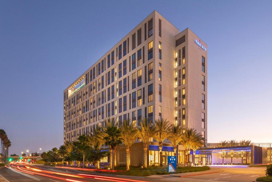 Modern multi-story hotel building with illuminated windows and palm trees in front. Evening sky sets the backdrop.