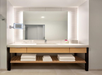 Bathroom vanity with a large mirror, a mounted towel rack below a countertop with a sink, toiletries, and a red flower in a vase. Folded towels are stored on a wooden shelf underneath.