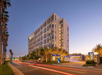 Modern multi-story hotel building with palm trees, lit by streetlights, and light trails from passing cars at dusk.