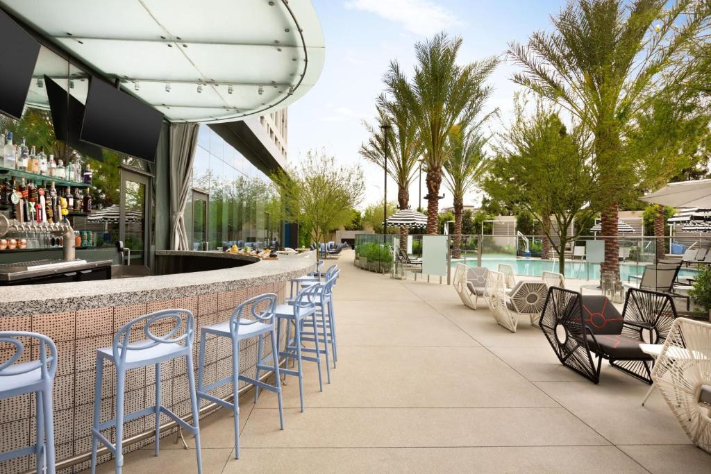 Outdoor bar area with blue stools, a curved counter, and a pool in the background. Palm trees and lounge chairs are visible under a partly cloudy sky.
