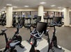 A gym room with stationary bikes in the foreground and treadmills with screens in the background. The room has beige walls and a low ceiling with overhead lighting.