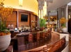 Modern bar with a curved counter, wooden stools, and a large yellow light fixture. Plants and bottles are displayed, and a TV is mounted above the counter.