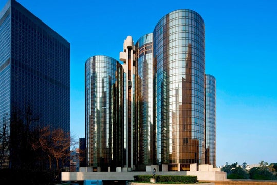A modern skyscraper with curved glass and steel design reflecting the blue sky, surrounded by trees and smaller buildings.