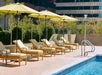 Poolside loungers with yellow umbrellas on a sunny day. Steps lead into the pool, surrounded by glass fencing and buildings in the background.