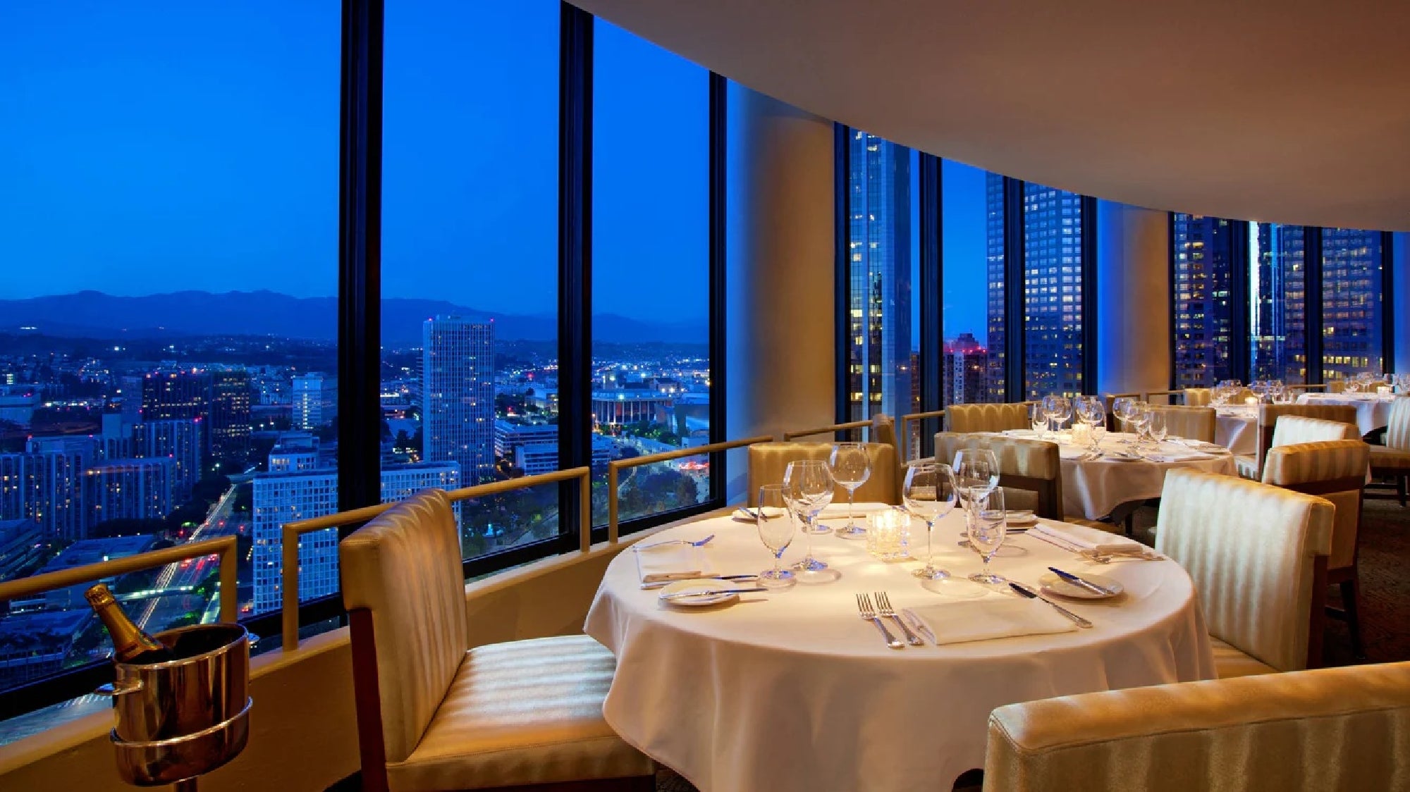 Elegant restaurant with panoramic cityscape view, round tables set with wine glasses and napkins, large floor-to-ceiling windows, and ambient evening lighting.